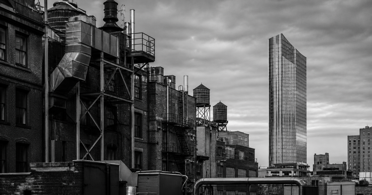 A dense industrial rooftop of brick, steel, ducts, and water towers contrasted with a single calm modern tower beneath an overcast sky.