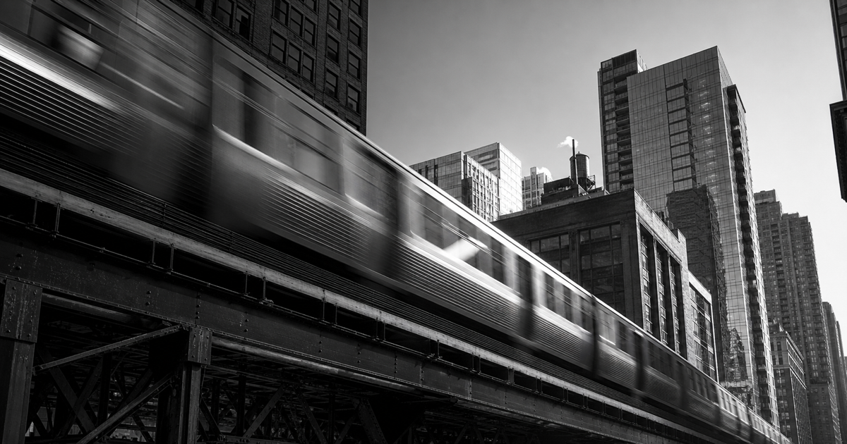 A commuter train streaks along elevated tracks between sharply rendered downtown buildings in black and white.